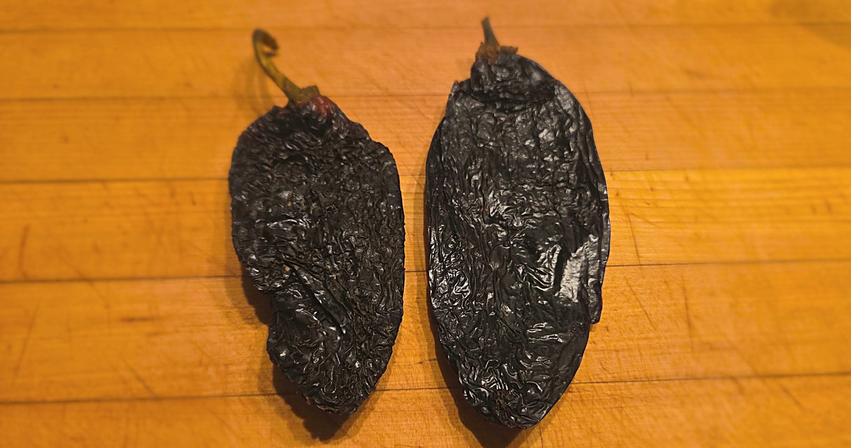 Two whole dried ancho chiles with wrinkled dark brown skin resting on a wooden cutting board